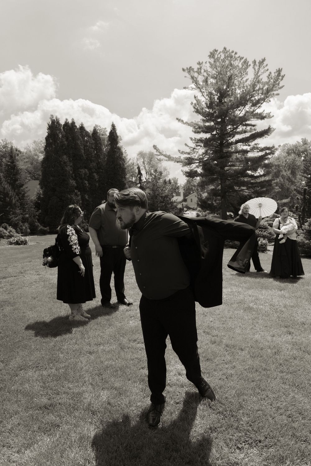 Black and white photo of wedding guests gathered on lawn with trees and cloudy sky backdrop.