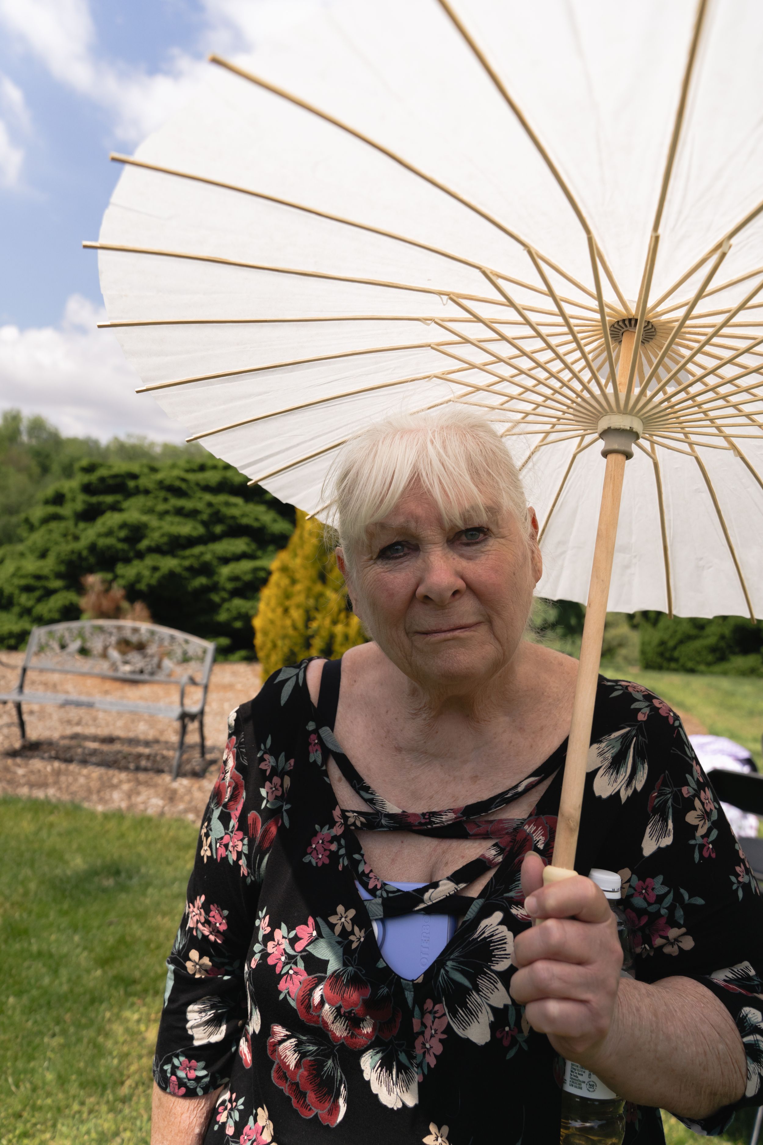 Elderly woman holding white parasol umbrella outdoors at a sunny garden wedding event.