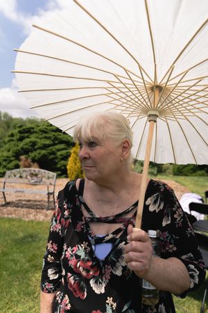 Elderly woman in floral dress holding white paper parasol umbrella outdoors at garden event.