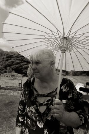 Elderly woman holding a white parasol umbrella outdoors at a garden event in black and white photography.