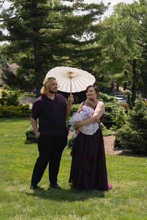Couple poses outdoors with white parasol, woman holds baby, man in purple shirt, lush garden background.