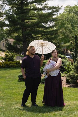 Couple in formal attire holding a white parasol and baby at an outdoor wedding celebration.