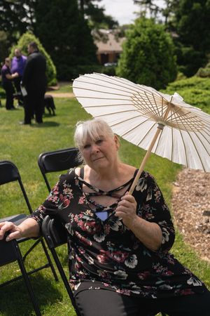 Elderly woman holding white parasol umbrella seated in black folding chair at outdoor wedding.