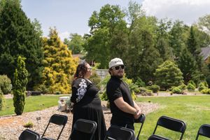 A couple stands back-to-back outdoors at a wedding venue surrounded by lush green trees and black folding chairs.