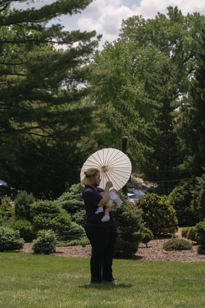 Woman holding baby under cream parasol umbrella in lush garden setting.