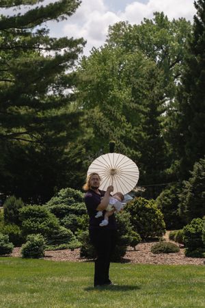 Woman in purple dress holding baby under white parasol in lush green garden landscape.