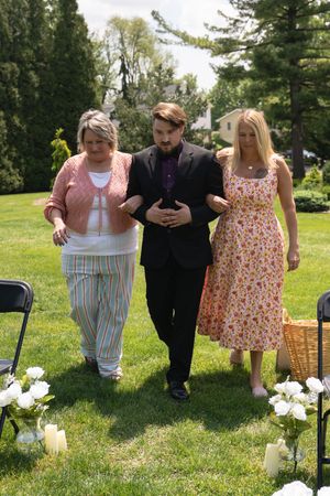 Groomsman in black suit escorts two women down outdoor wedding aisle lined with white flowers.