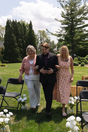 Groom escorted down the aisle by two women at an outdoor garden wedding ceremony on a sunny day.