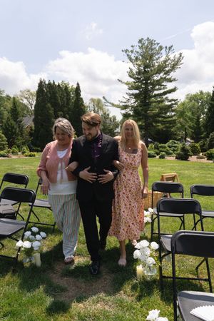 Groomsman walks two female guests down a sunlit outdoor wedding aisle with black folding chairs.