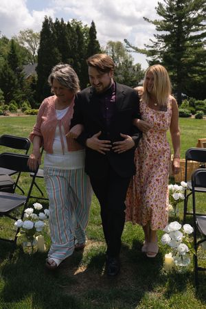 Groom escorted by two women down outdoor wedding aisle lined with white floral arrangements.