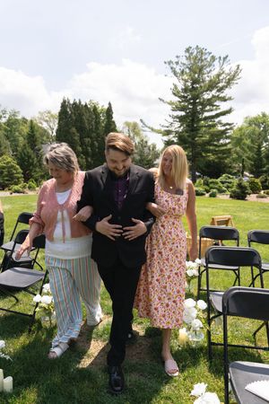Groom in black suit escorted down outdoor wedding aisle by two women in a garden ceremony.
