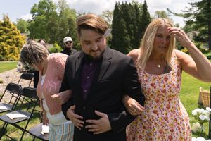 Man in purple suit escorted by two women in floral dresses at outdoor wedding ceremony.