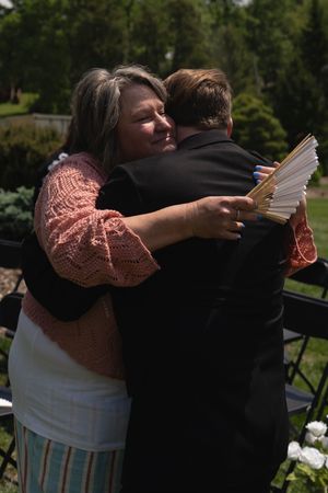Woman in pink cardigan hugging a man in a black suit at an outdoor wedding ceremony.