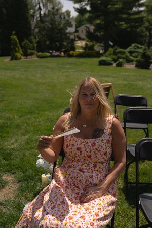 Blonde woman in floral dress fanning herself at outdoor wedding ceremony.