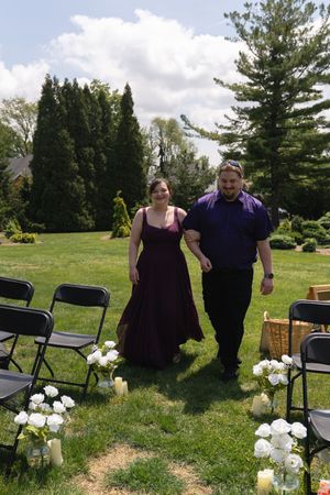 Couple in purple and burgundy walking down outdoor wedding aisle lined with white flowers.