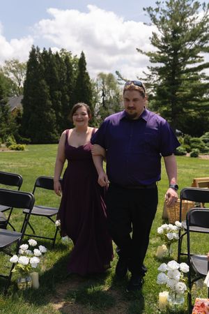 Bridesmaid in purple gown walks down outdoor wedding aisle with groomsman in black pants.