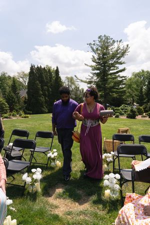 A man in purple and woman in a pink dress walk down an outdoor wedding aisle decorated with white flowers.