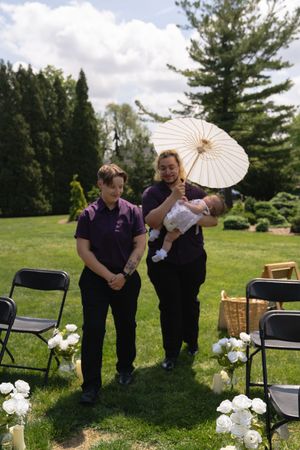Two people in purple shirts walking down wedding aisle, one holding baby and white parasol.