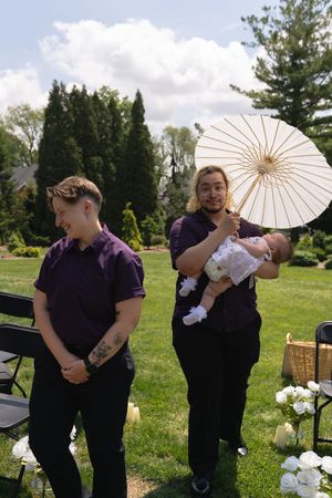 Two groomsmen in purple shirts at outdoor wedding, one holding parasol and newborn baby.