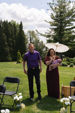 Man in purple shirt and woman in burgundy gown holding baby and parasol at outdoor wedding.