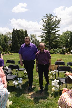Two men in purple shirts walk down outdoor wedding aisle lined with white floral arrangements.