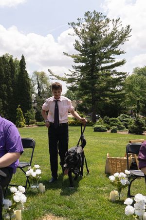 Young man in formal wear walks a black dog down a flower-lined outdoor wedding ceremony aisle.