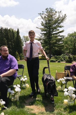 Young man in pink shirt and tie walking a black dog down the aisle at an outdoor garden wedding.