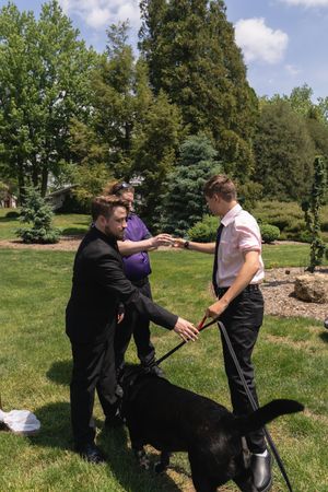 Two men greeting each other with a handshake while a black dog stands nearby in a garden.