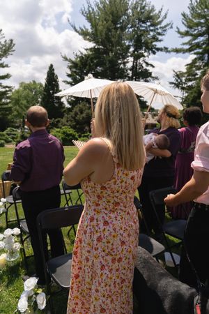 Outdoor wedding ceremony guests standing, woman in floral dress watching procession with parasol.