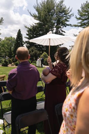 Wedding guests seated outdoors watching a ceremony, woman holding baby under white parasol umbrella.