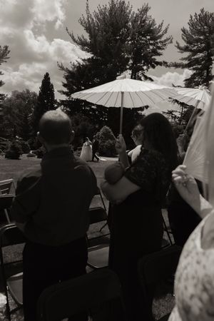 Guests seated outdoors at a wedding ceremony, one holding a white parasol, with tall trees in the background.