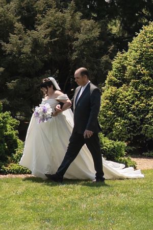 Bride in white ball gown holding purple bouquet walks with escort through a garden wedding.