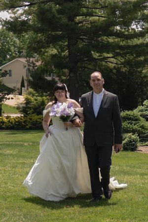 Bride in white gown holding purple bouquet walks with escort across green lawn at outdoor wedding.