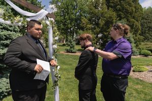 Officiant reads vows while groomsman adjusts boutonniere on groom at outdoor wedding ceremony.