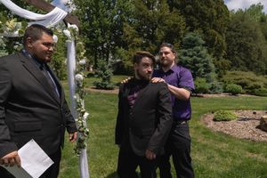Groom receiving emotional support from groomsmen near floral arch before outdoor wedding ceremony.
