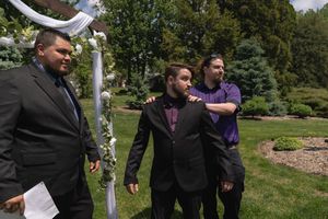 Three men stand under floral arch at outdoor wedding ceremony, one holding papers.