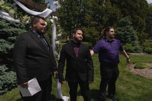 Three men in formal attire standing near floral wedding arch in outdoor garden ceremony.