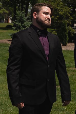 Man in black suit with purple shirt and bow tie standing outdoors in a garden setting.