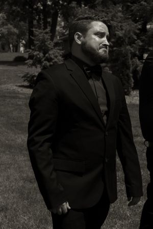 A black and white portrait of a man in a suit and bow tie standing outdoors among trees.