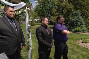 Groom and two men stand at floral wedding arch during outdoor garden ceremony on sunny day.