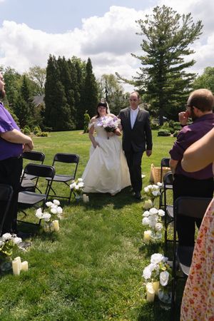 Bride in white gown walks down outdoor aisle with escort past seated guests and white flowers.