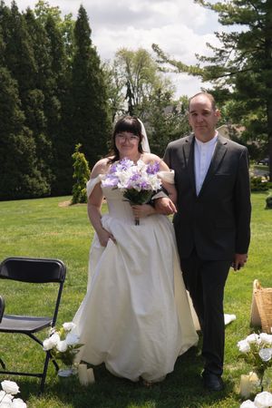 Bride in white gown holding purple bouquet walking aisle with escort.