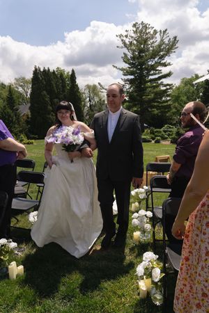 Bride in white gown with bouquet walks aisle with escort at outdoor garden wedding ceremony.