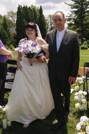 Bride in white gown holding purple and white bouquet walking down aisle with escort at outdoor wedding.