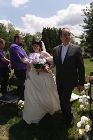 Bride in white gown holding purple bouquet walks down outdoor aisle with escort at garden wedding.