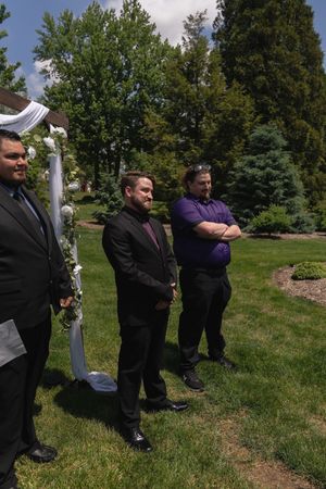 Three men in formal attire standing near floral wedding arch during outdoor ceremony on sunny day.