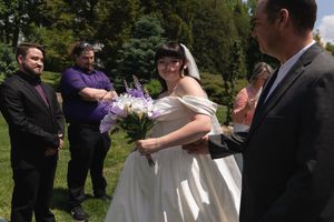Bride in white gown holding purple bouquet smiles at groom during outdoor wedding ceremony with guests nearby.