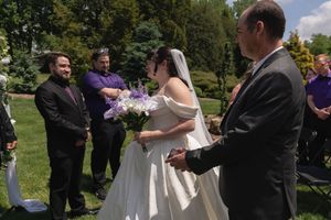 Father presenting bride to groom at outdoor wedding ceremony with groomsmen standing nearby.