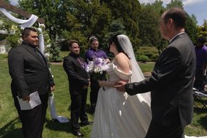 Bride in white gown with purple bouquet exchanges vows with officiant at outdoor garden wedding ceremony.