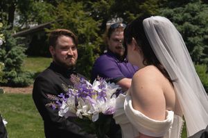 Groom smiling at bride holding purple and white bouquet during outdoor garden wedding ceremony.